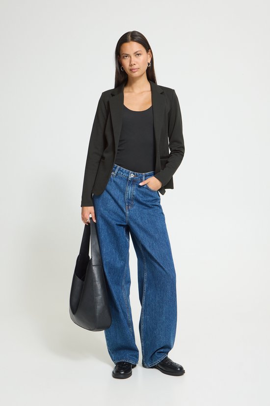 Model met lange zwarte blazer, één knoop sluiting, gedragen met een zwarte top, jeans en zwarte tas.