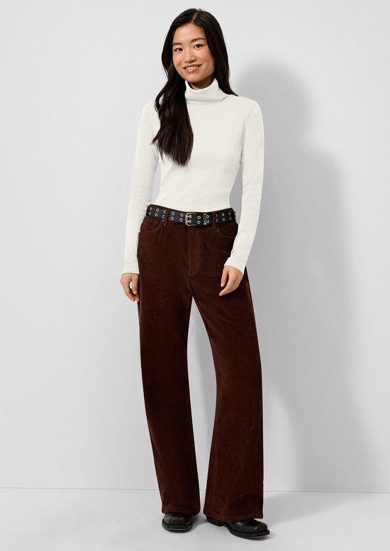 Model draagt een witte coltrui en bruine corduroy broek met riem.