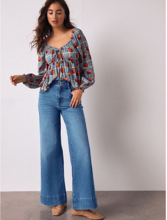 Model in een blauwe top met appelprint en gele ruit, met een hoge taille en wijde jeans.