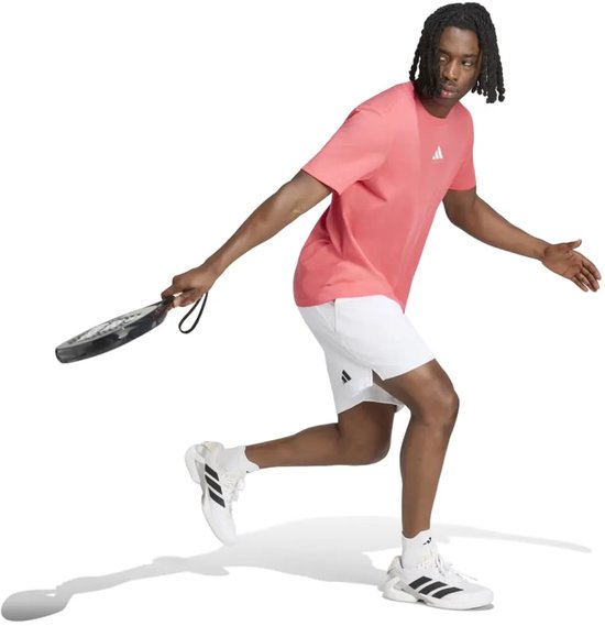 Man in een roze Adidas T-shirt en witte korte broek met een padelracket.