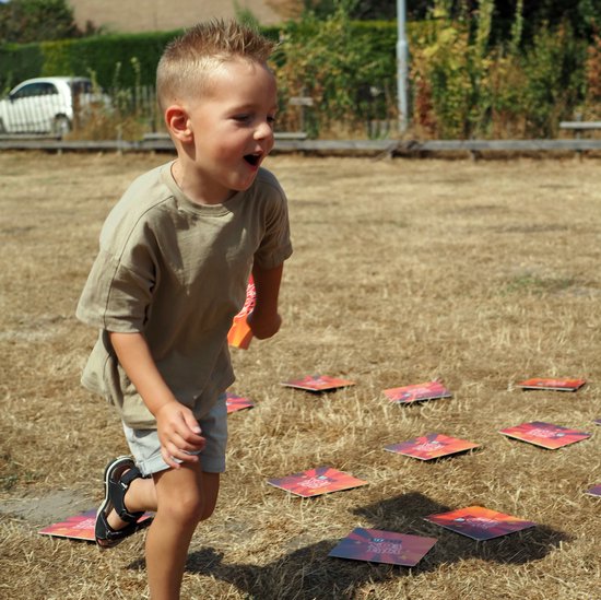 Jongen in beige shirt rent over droog gras, langs 'KIDS BOX PLAY XXL' kaarten.