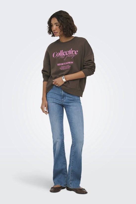 Vrouw staat in een bruine sweater met roze tekst en jeans.