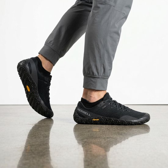 Man die Merrell Vapor Glove 6 zwarte sportschoenen draagt, met een close-up van de schoenen en een deel van zijn grijze broek.