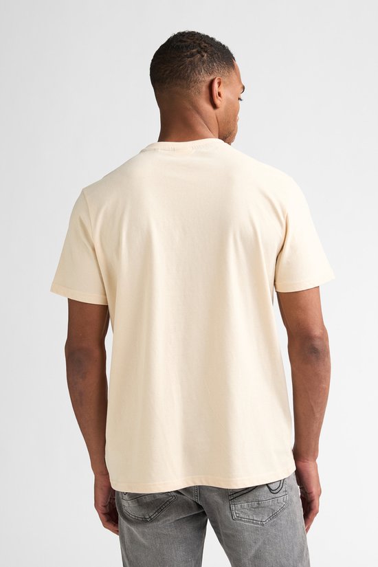 Man staat met de rug naar de camera, draagt een crème T-shirt met korte mouwen en grijze jeans.