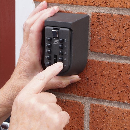 Hand die een zwarte metalen sleutelkluis op baksteen bedient. Vinger drukt cijfer 9 in.
