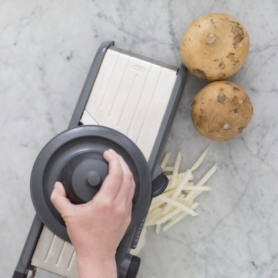 Een mandoline met antislipvoetjes snijdt aardappels in dunne reepjes. Een hand met ring draait de voedselhouder.