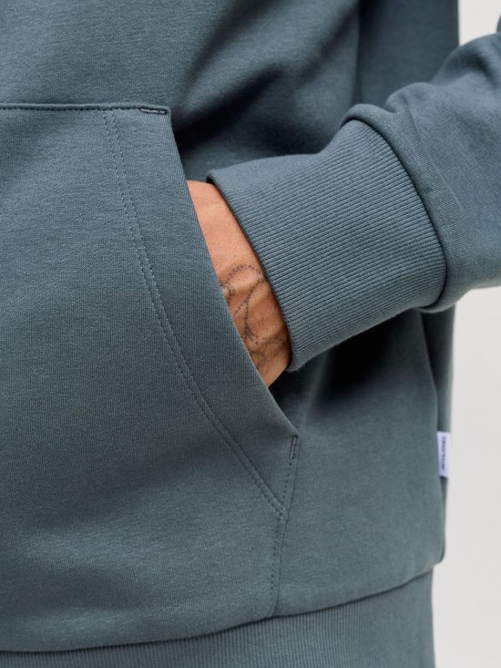 Gedetailleerde opname van een blauwgrijze hoodie met een kangoeroezak. Een hand met tatoeage steekt in de zak.