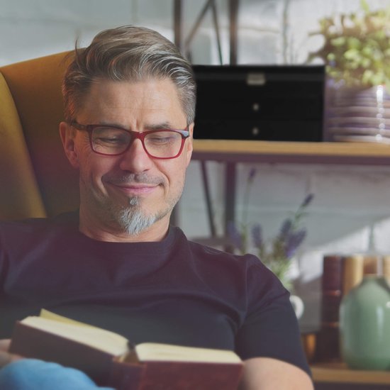 Man met bril en grijze baard leest een boek in een fauteuil.