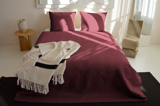Bordeauxrode sprei met gebreid kabelpatroon, twee kussenslopen, een plaid met franjes en gestreepte kussens op een bed.