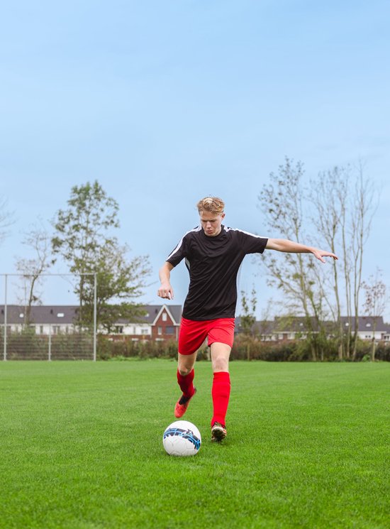 Jonge man in zwart voetbalshirt en rood voetbalbroekje trapt tegen een voetbal.