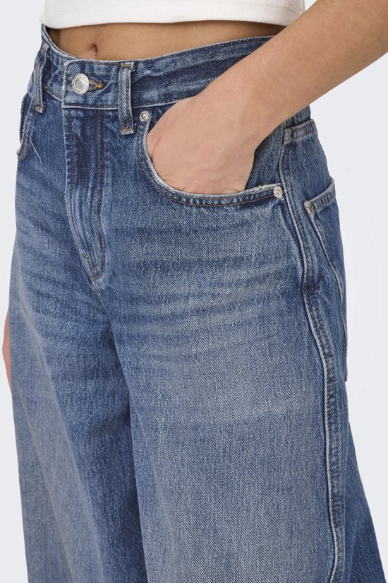 Close-up van een middendonkere, wijde spijkerbroek met hoge taille. Model steekt hand in de zak.