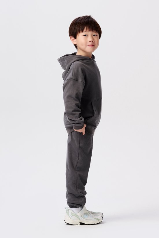 Jongen in donkergrijze hoodie met logopatch, joggingbroek en witte sneakers. Zijaanzicht.