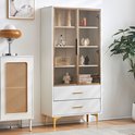Witte vitrinekast met glazen deuren, gouden accenten, gouden handgrepen en gouden poten. Gedecoreerd met beeldjes, een radio en boeken.