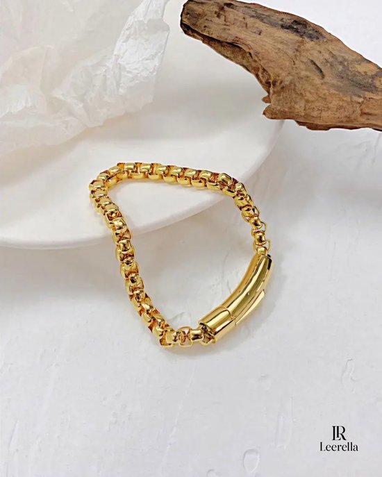 Gouden schakelarmband met gesp op een witte ondergrond, met een stuk drijfhout.