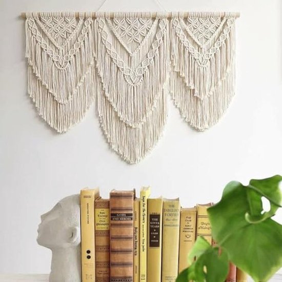 Crèmekleurig macramé wandkleed met drie V-vormige patronen, boven een rij boeken en een beeld.