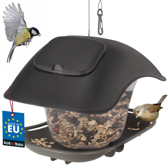 Grijs vogelvoederhuisje met transparante silo en donkergrijs dak, gevuld met vogelzaad. Een grote mees vliegt weg, een kleine mees zit op de rand. Blauw 'Made in EU' label.