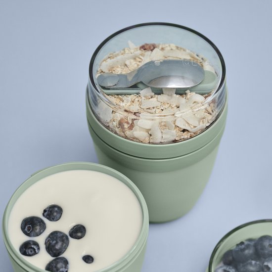 Lunchpot vert avec couvercle transparent, rempli de muesli et de copeaux de noix de coco. À côté du pot, un récipient avec du yaourt et des myrtilles.
