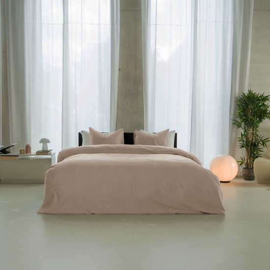 Een compleet opgemaakt bed met oudroze beddengoed, in een slaapkamer met grote witte gordijnen.