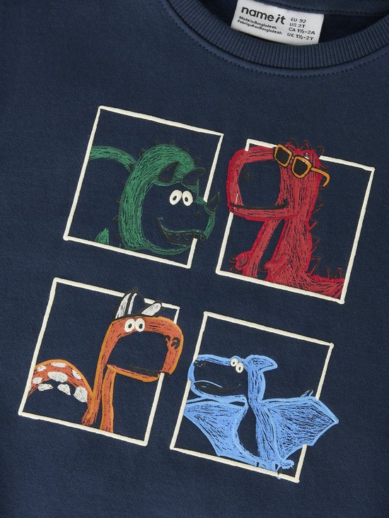 Close-up van de print op een donkerblauwe sweater: vier dinosaurussen in vakjes. Groene, rode met zonnebril, oranje en blauwe.