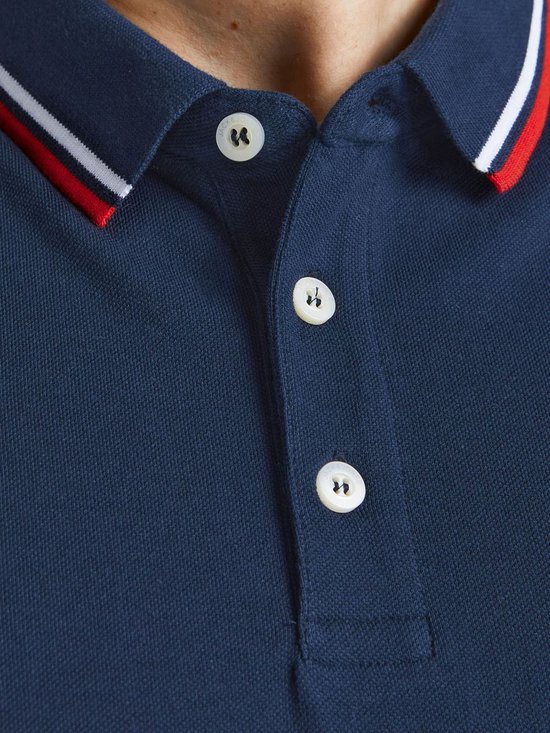 Man met een donkerblauwe polo met rode en witte strepen aan kraag en mouwen, lachend.