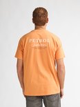 Man van achteren draagt een oranje t-shirt met "PETROL Industries" op de rug en een grijze jeans.