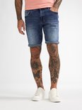 Man, staand, in een roze T-shirt en spijkerbroek met opgerolde zoom, witte sneakers.