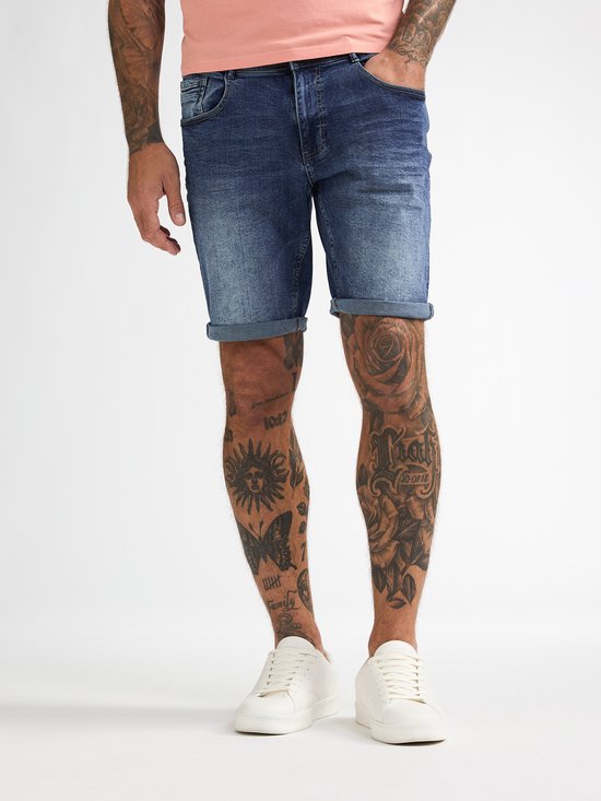 Man, staand, in een roze T-shirt en spijkerbroek met opgerolde zoom, witte sneakers.