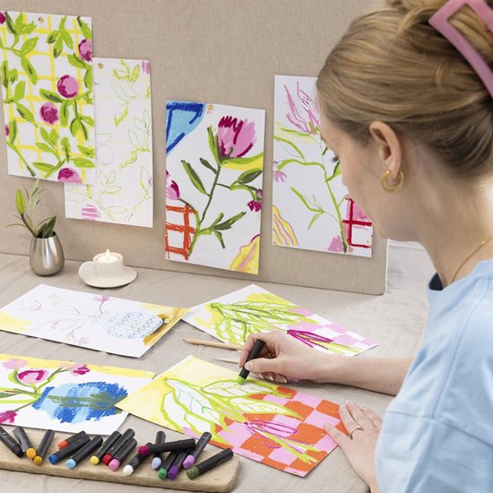 Een vrouw tekent bloemen met groene en oranje oliepastels op een geruit vel papier. Diverse schetsen en kleurrijke pastels liggen op tafel.