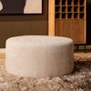 Ronde, stoffen poef in de kleur beige, geplaatst op een hoogpolig vloerkleed.