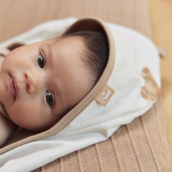 Le visage d'un bébé allongé dans une cape de bain avec un imprimé d'ours brun. La cape de bain a un bord et une étiquette beiges.