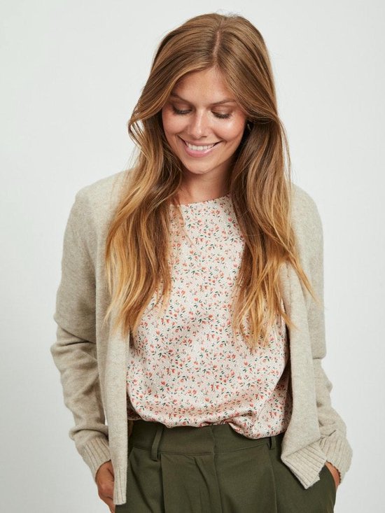 Een lachende vrouw met lang blond haar, draagt een beige vest over een bloemenblouse.