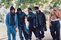 Vijf mannen lopen buiten in een groep, gekleed in casual kleding en denim jacks. Eén man is zonder shirt.