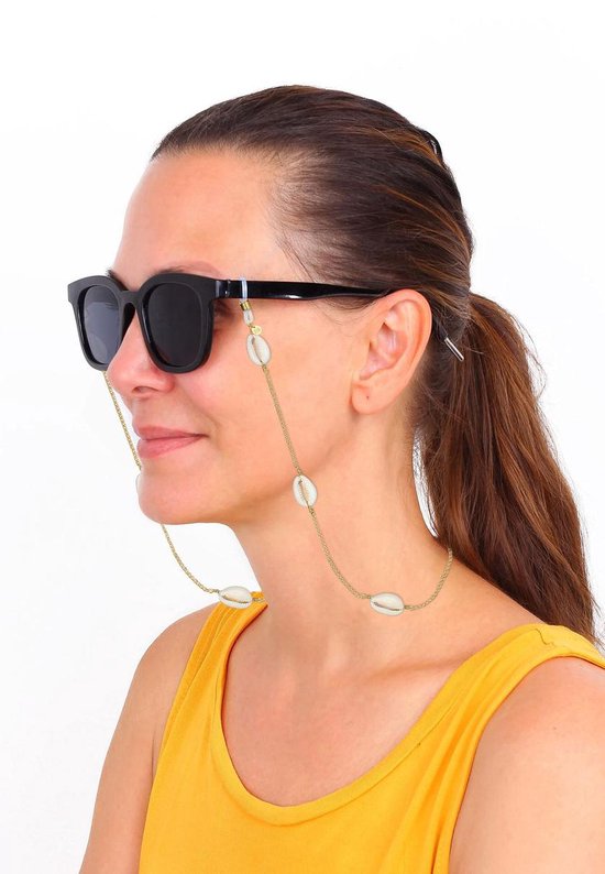 Model met zwarte zonnebril en een gouden brilketting met kauri schelpen en oranje-gele kralen.