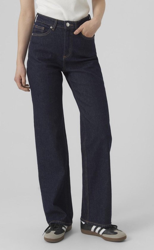 Model draagt donkerblauwe wijde jeans met oranje stiksels, een witte top en sneakers.