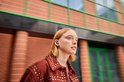 Een blonde vrouw met een bruin gespeld jasje draagt een donkergrijs oordopje.