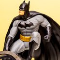 Batman-figuur in grijs pak met gele riem en zwarte cape, zittend op een lamp. Een donkere achtergrond.