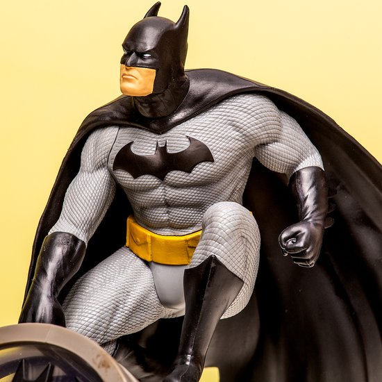 Batman-figuur in grijs pak met gele riem en zwarte cape, zittend op een lamp. Een donkere achtergrond.
