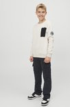 Jongen in gebroken witte sweater met zwarte borstzak en ronde patch op mouw, en donkergrijze cargo broek.
